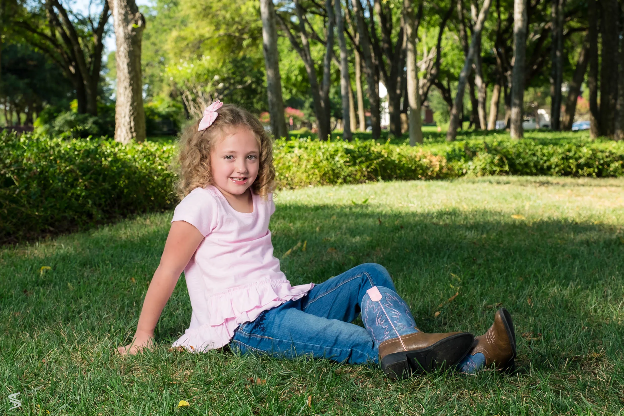 Young girl in a pink shirt and cowboy boots sitting on the grass beneath shady park trees.