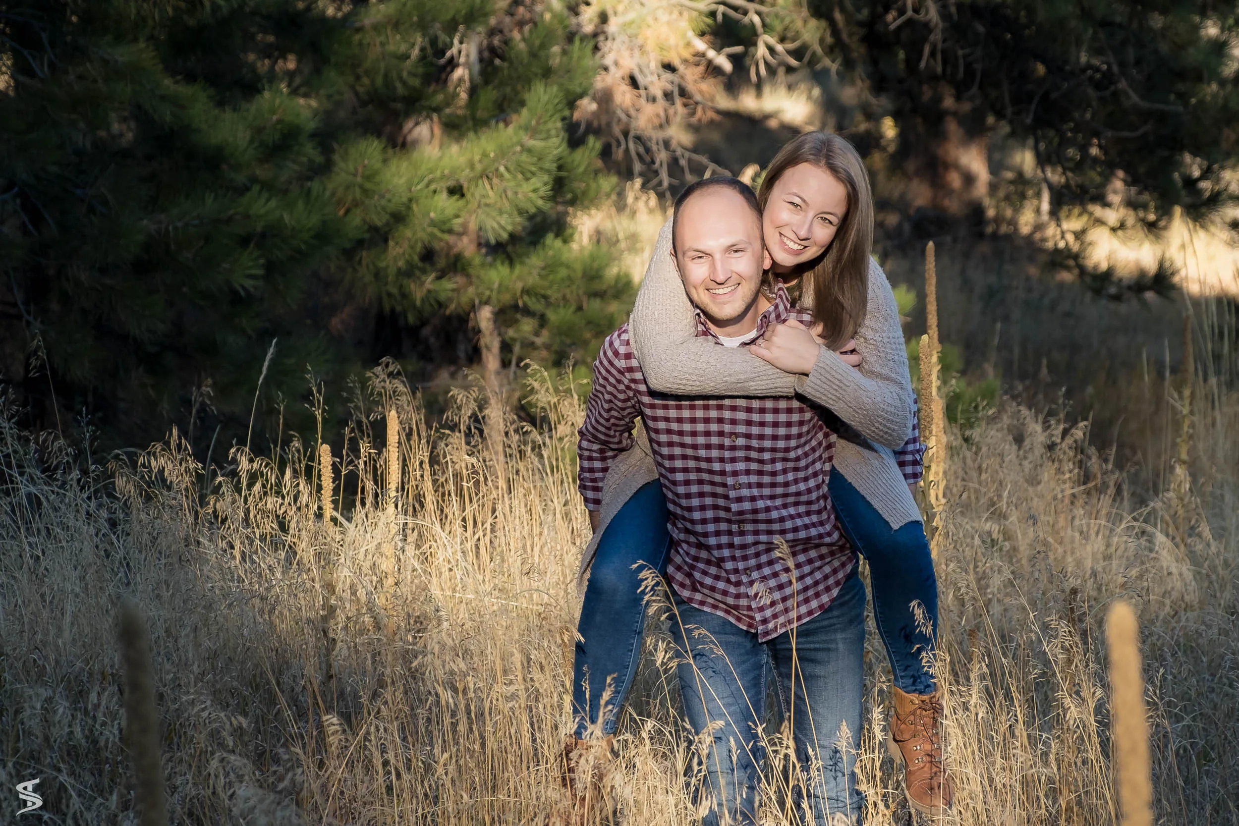 Smiling woman riding piggyback on her partner through tall grass beside pine trees.