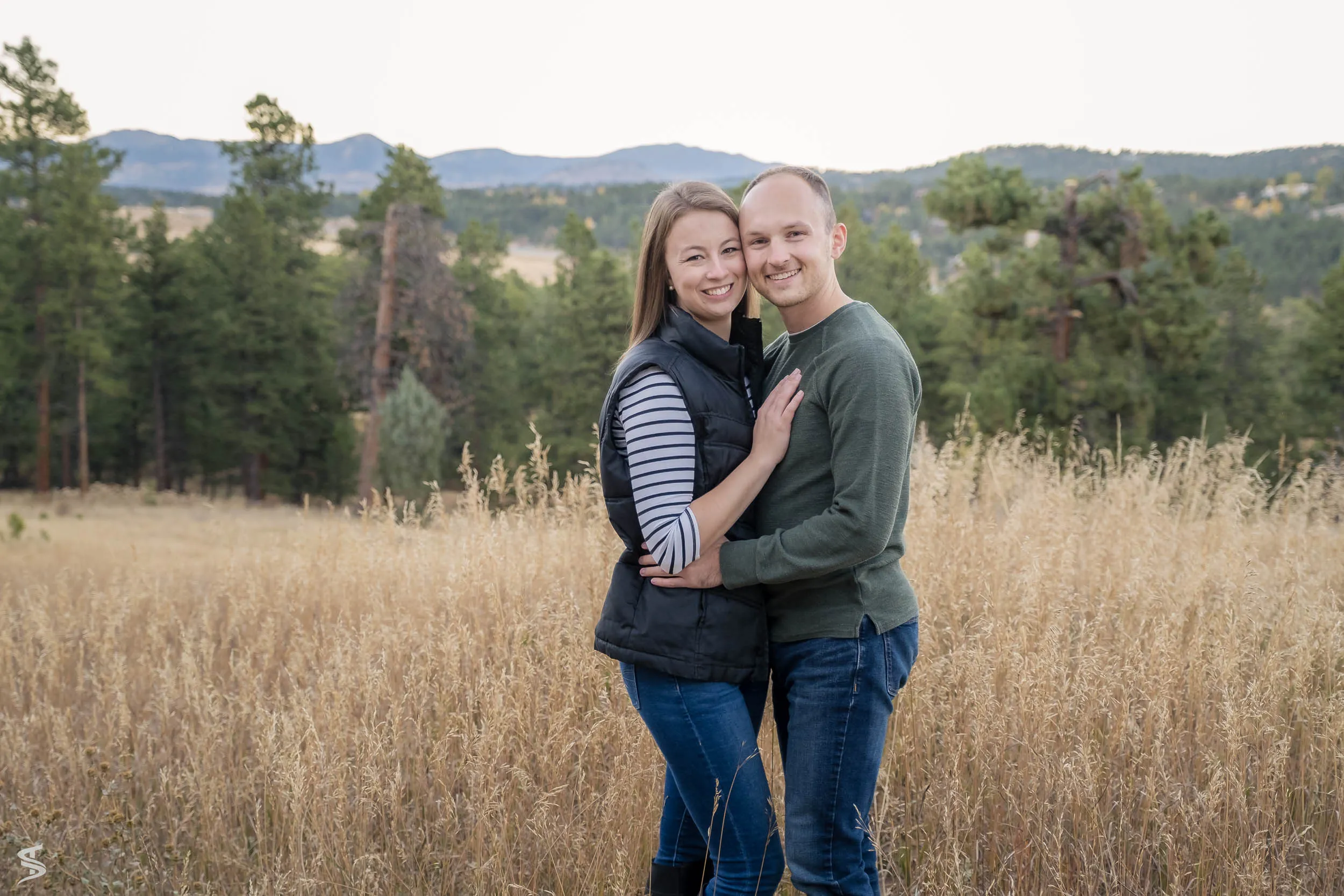 Couple embracing in a grassy meadow with pine trees and distant mountains behind them.