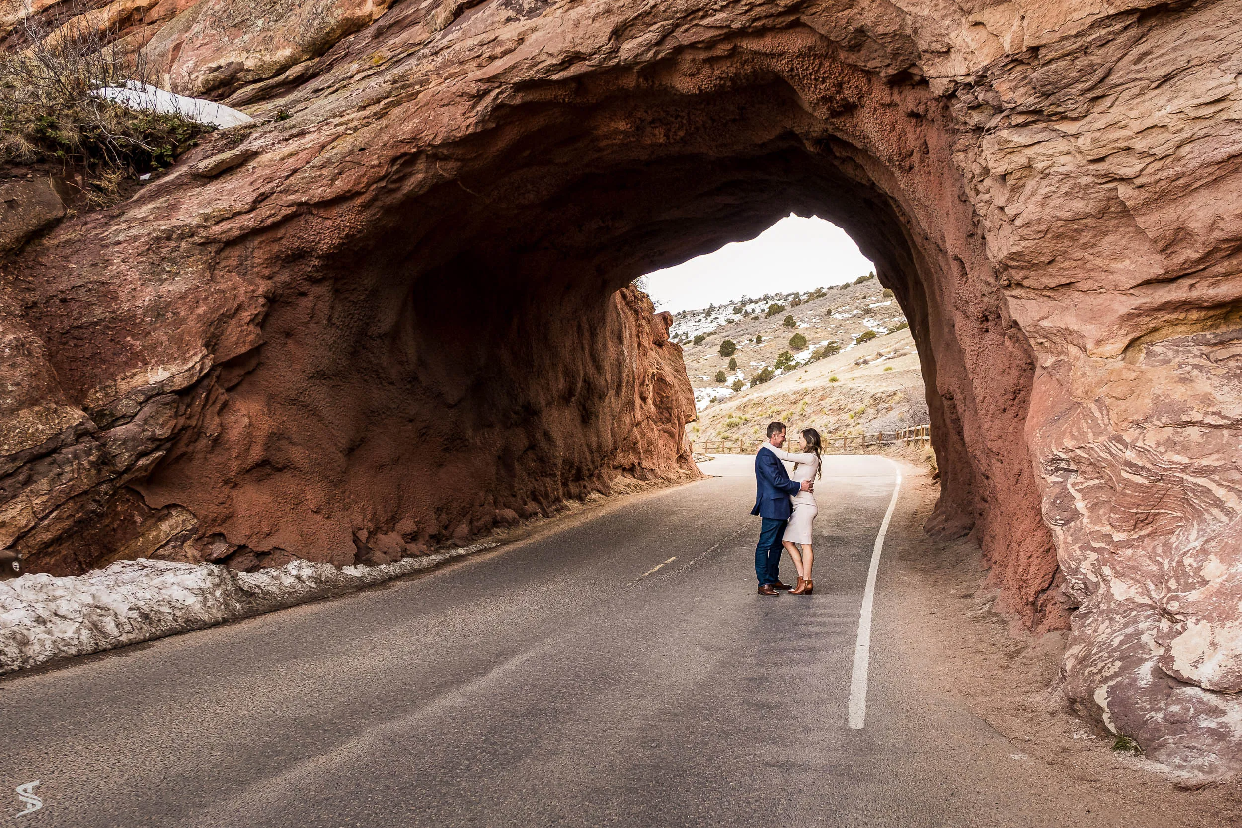 Couple embracing on a winding road beneath a natural red rock arch.
