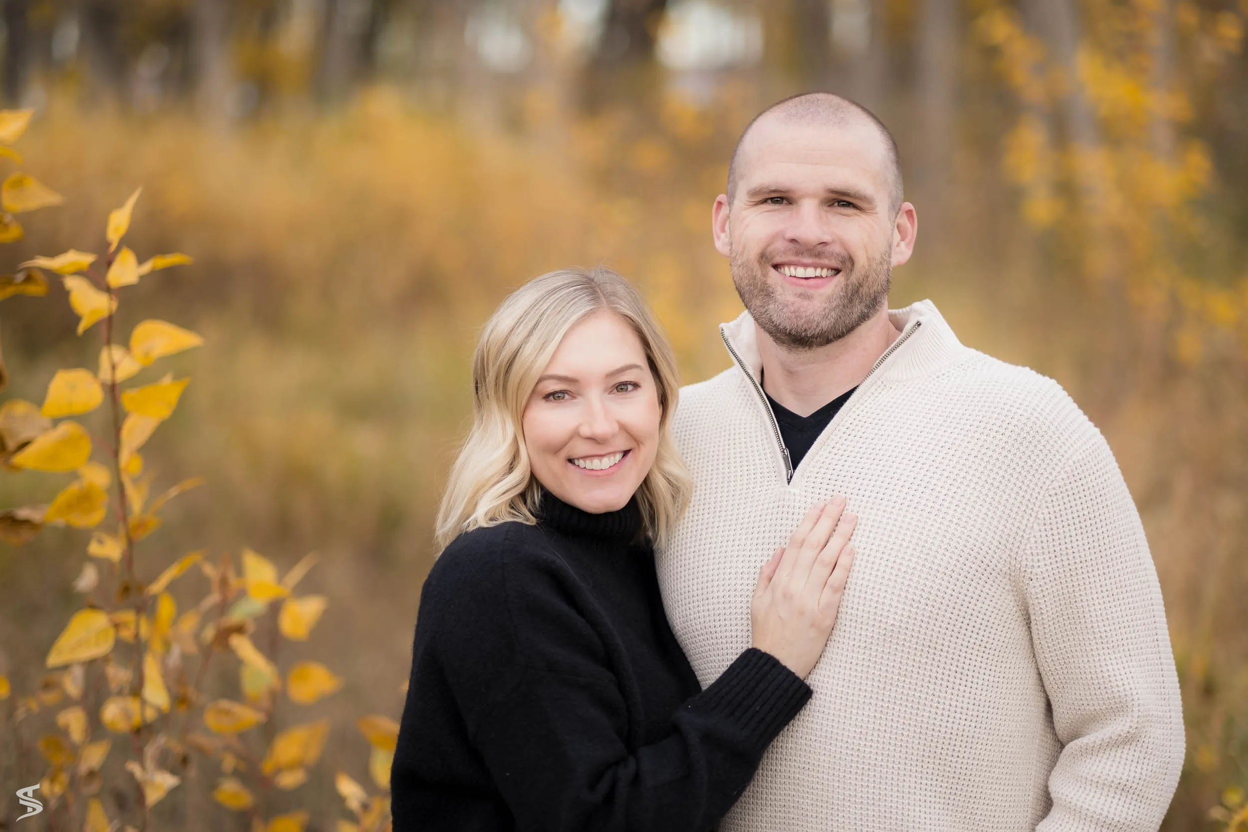 Close-up portrait of a smiling couple surrounded by golden autumn leaves.