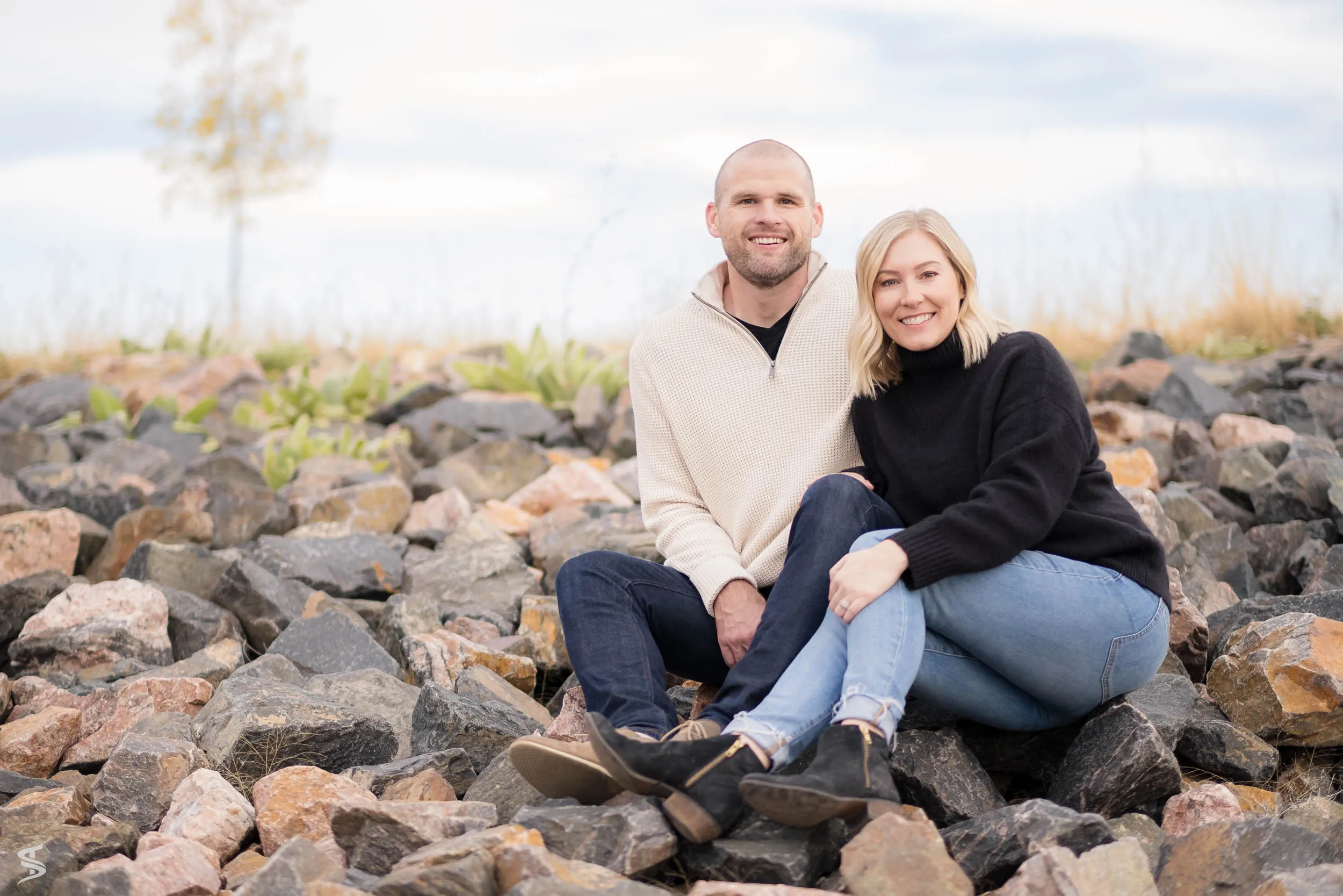 Couple sitting close together on a rocky shoreline beneath a cloudy sky.