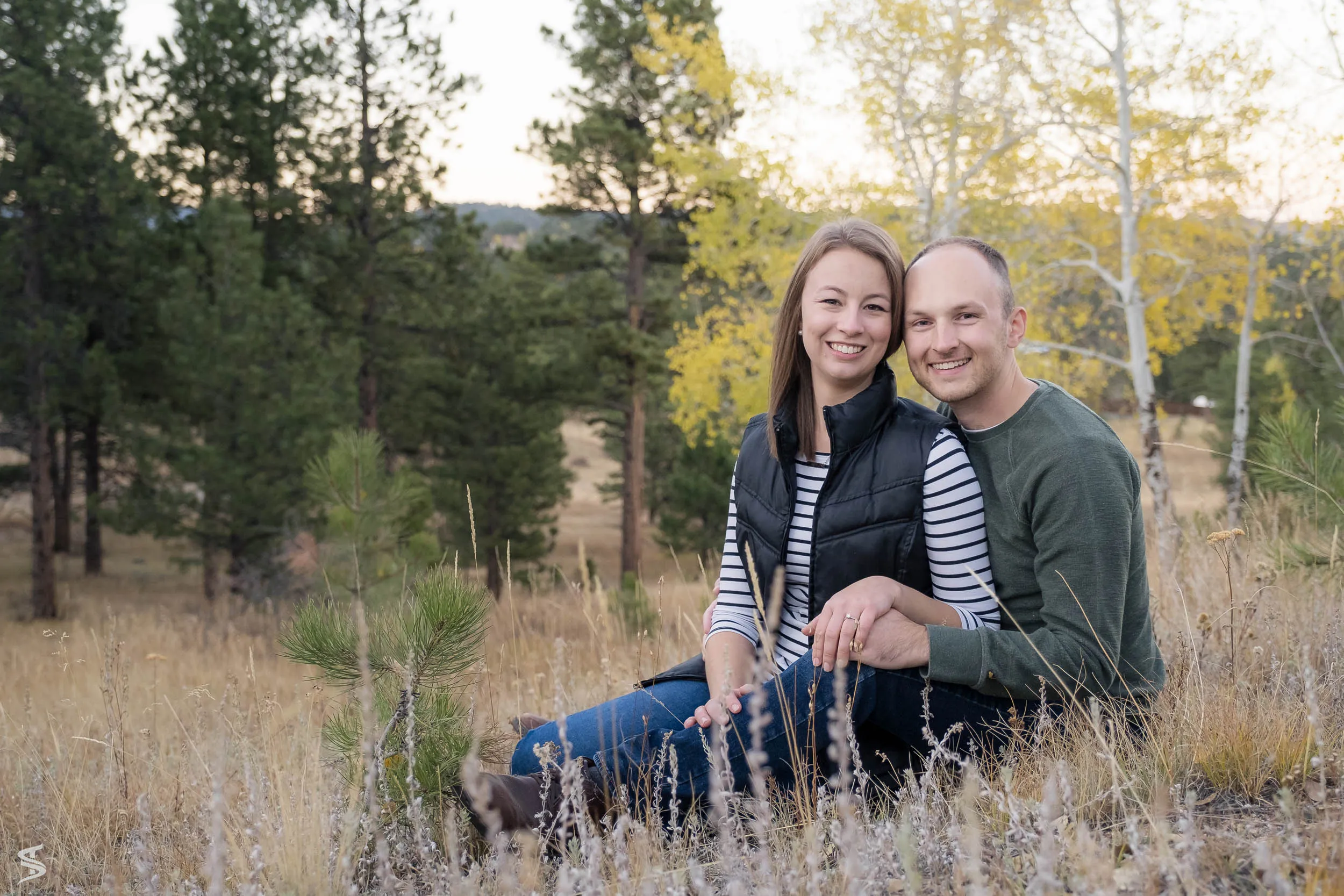 Smiling couple seated together among tall grasses, with autumn foliage and pine trees behind them.