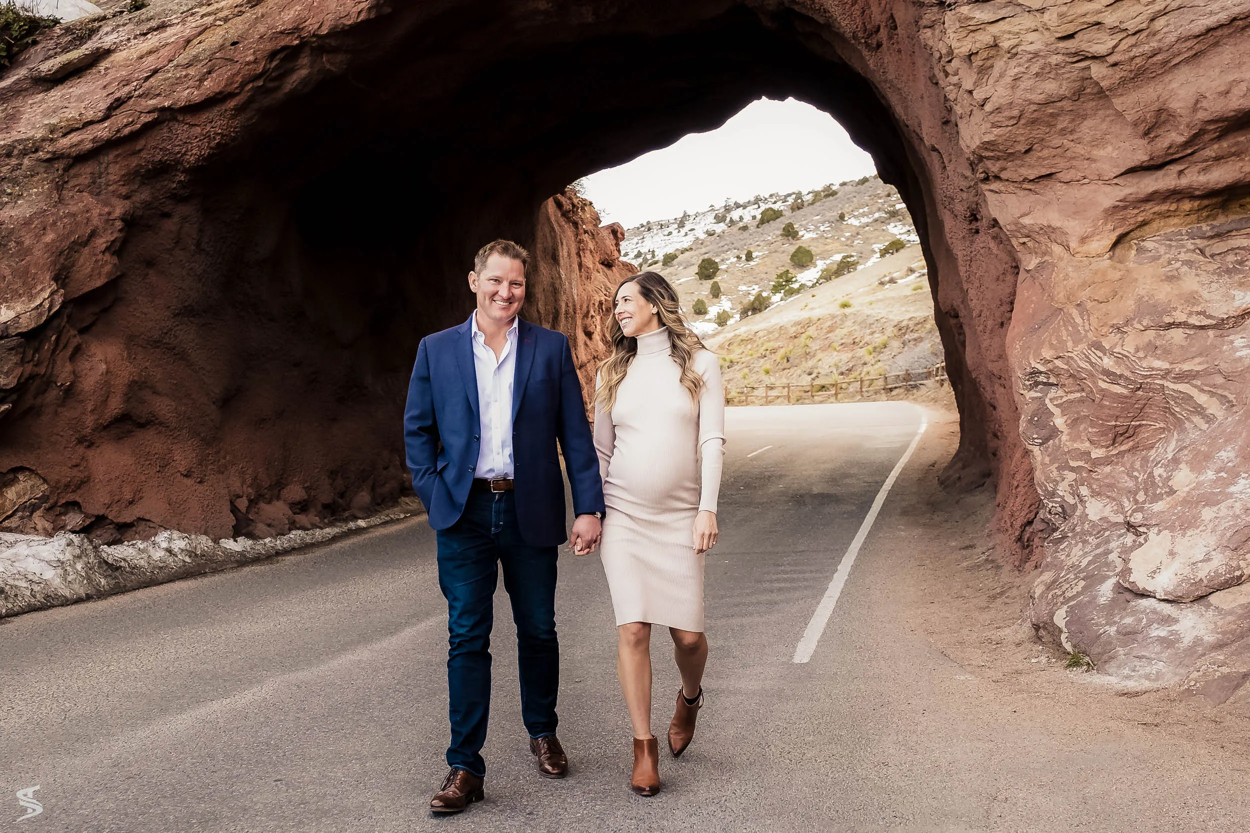 Couple walking hand in hand along a road beneath a red rock arch.