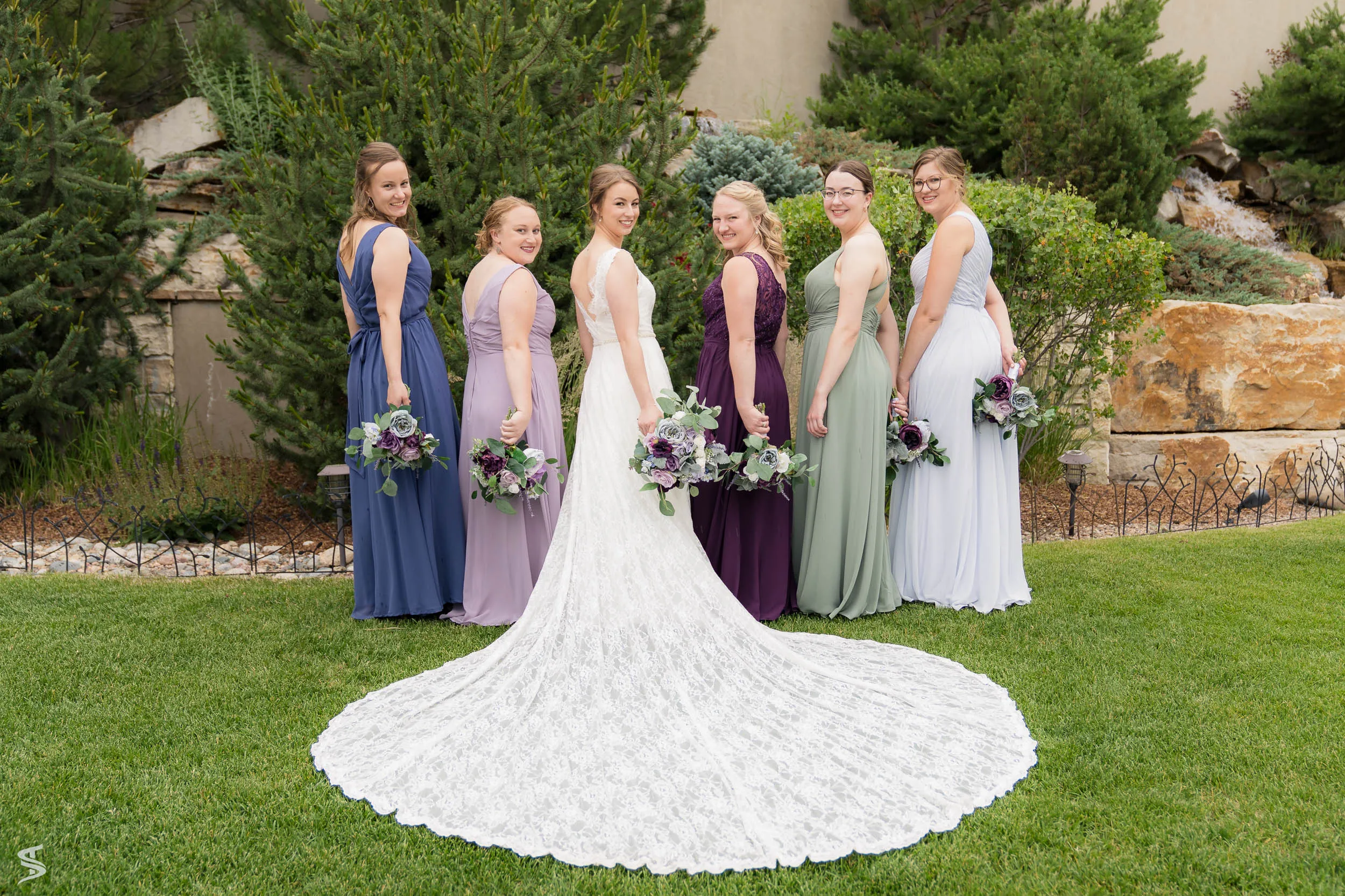 Bride posing with five bridesmaids in colorful dresses, with her long lace train spread across the lawn.