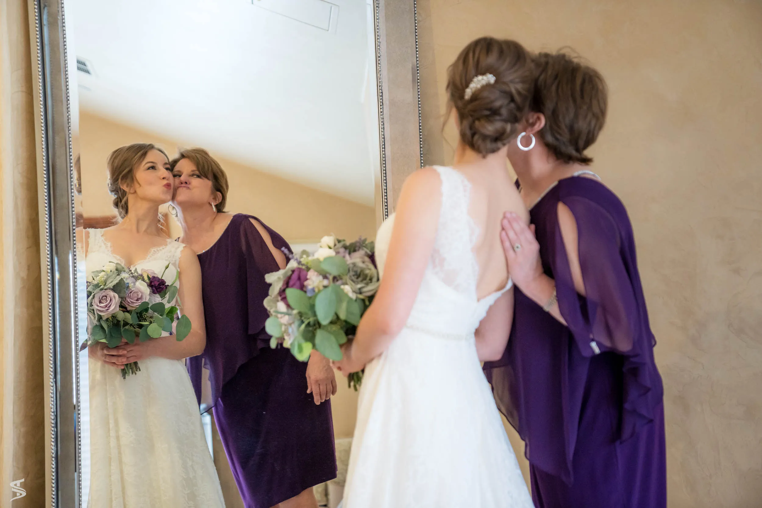 Woman in a purple dress kissing the bride on the cheek, with their reflection visible in a mirror.