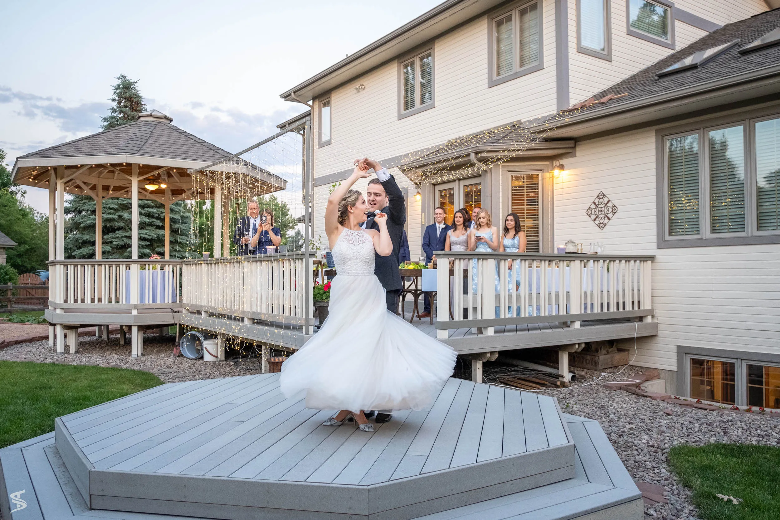 Groom twirling the bride on an outdoor wooden dance floor while wedding guests watch from the deck.