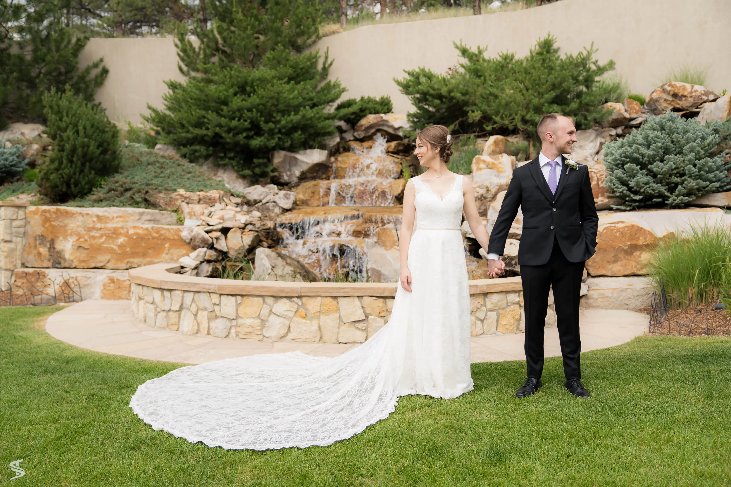 Bride and groom holding hands on a garden lawn beside a cascading rock waterfall, with her lace train spread behind her.