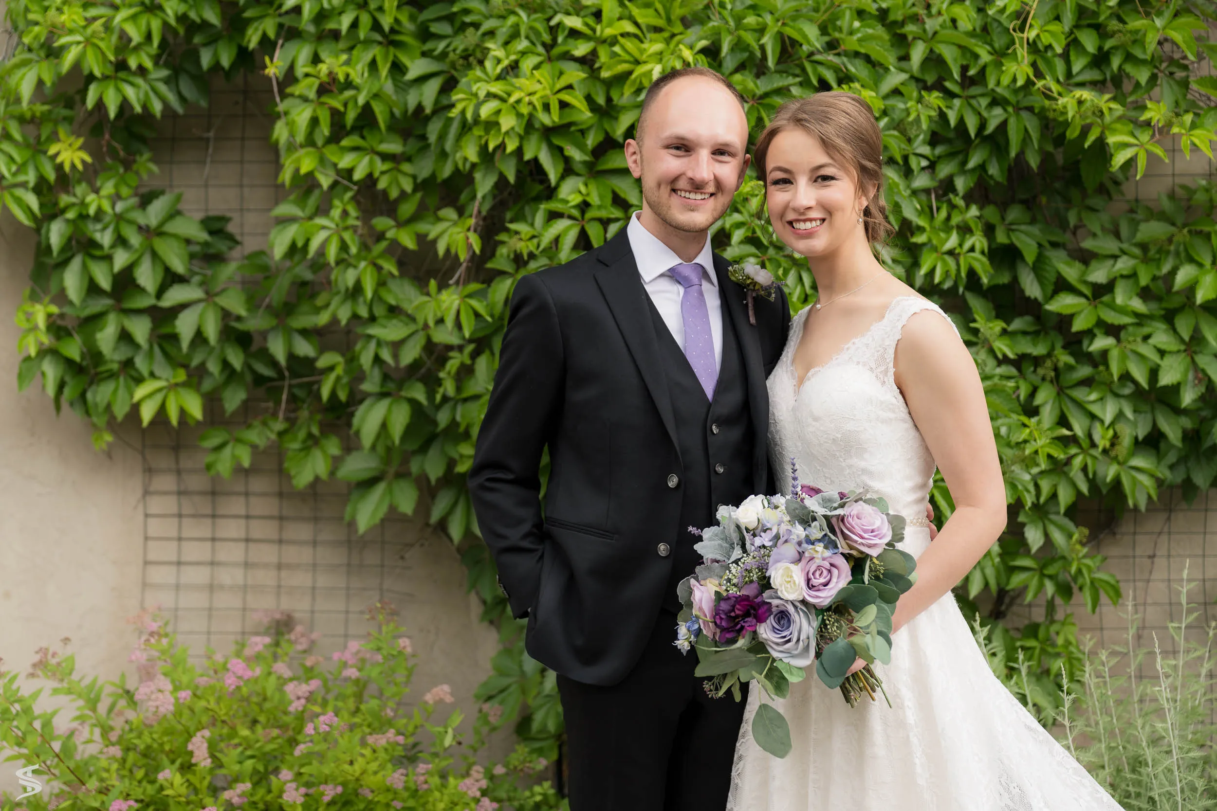 Smiling bride and groom posing against a leafy garden wall, with the bride holding a purple and white bouquet.
