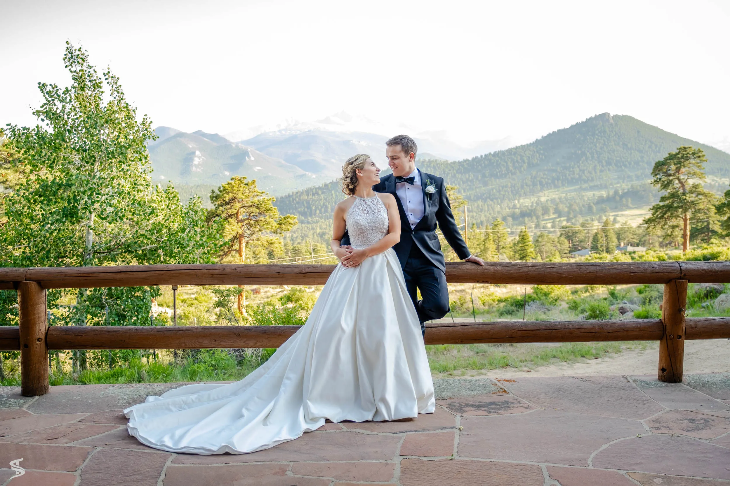 Bride and groom standing beside a wooden railing on a stone terrace overlooking mountains.
