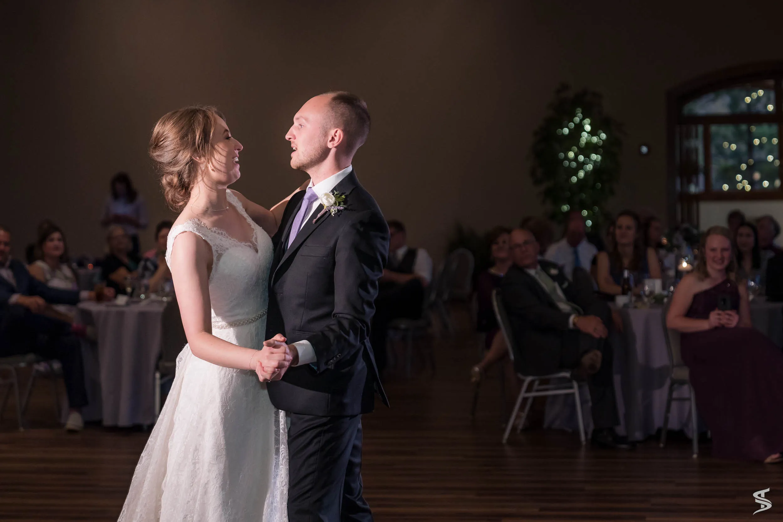 Bride and groom smiling at each other while dancing together, with wedding guests seated around the reception floor.