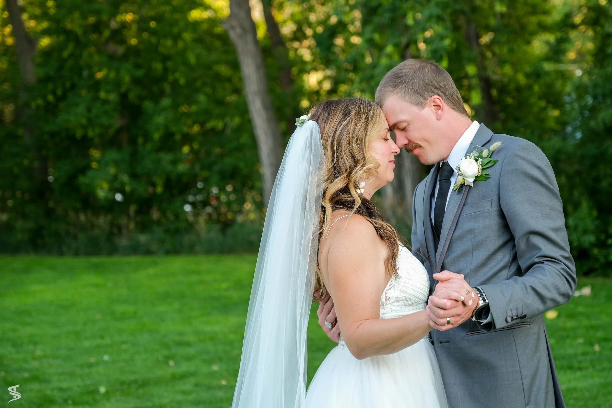 Bride and groom touching foreheads and holding hands during a quiet wedding portrait in a green garden.