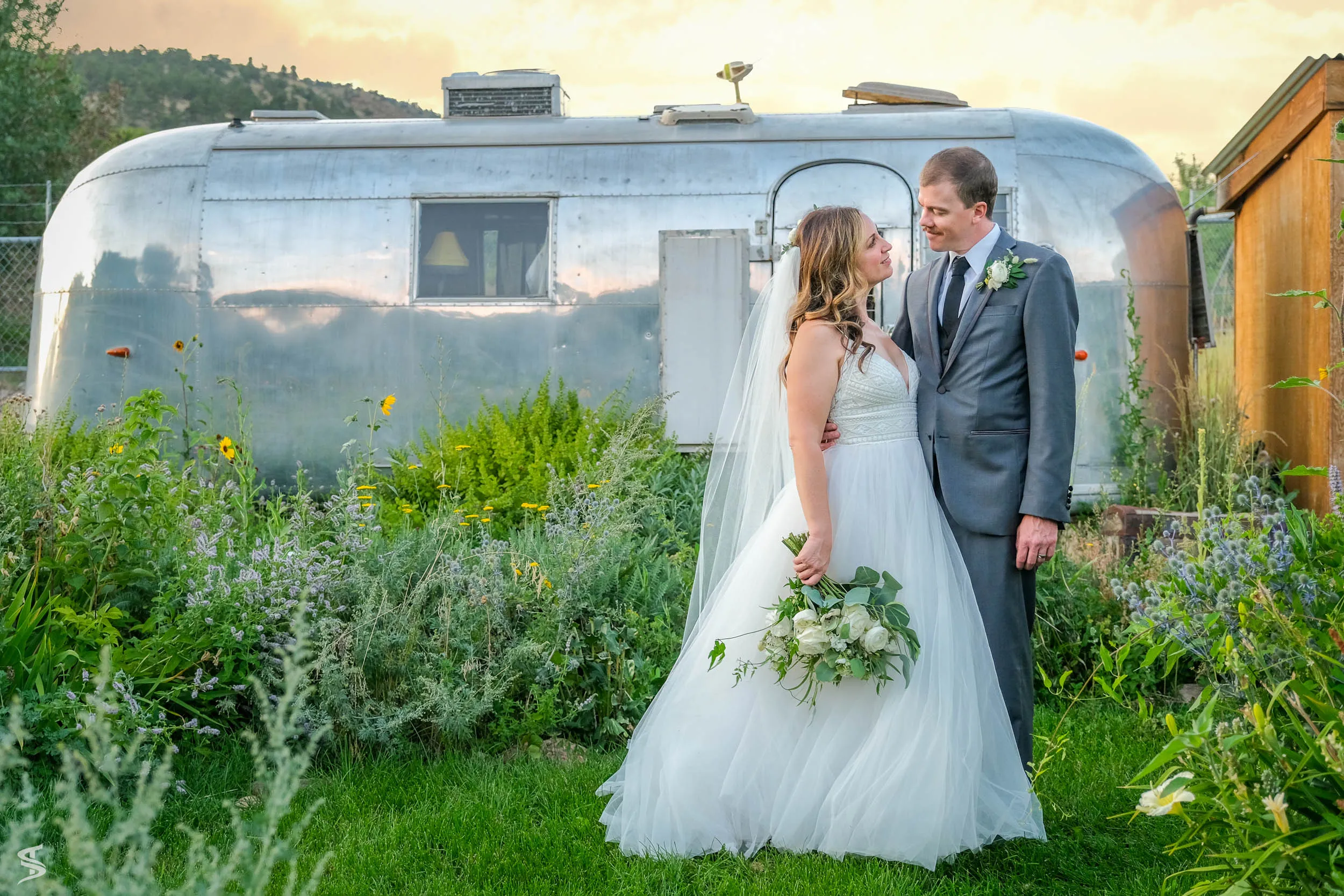 Bride and groom standing together beside a vintage silver travel trailer surrounded by garden flowers.