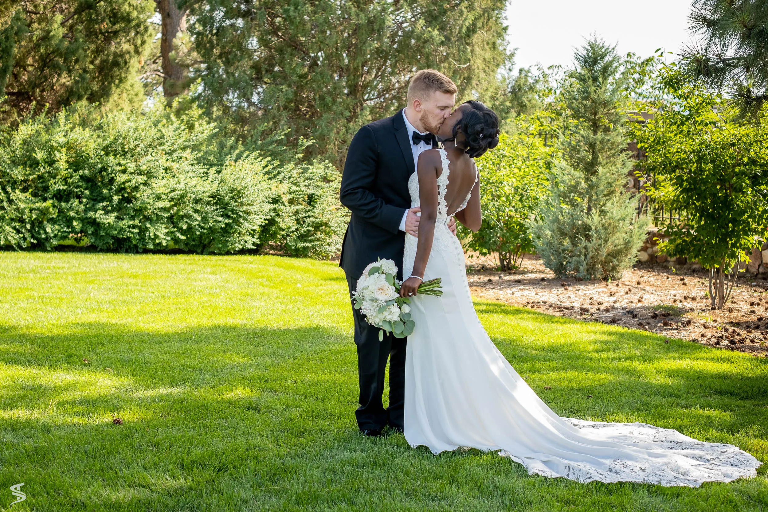 Bride and groom kissing in a sunlit garden, with her lace dress train spread across the lawn.