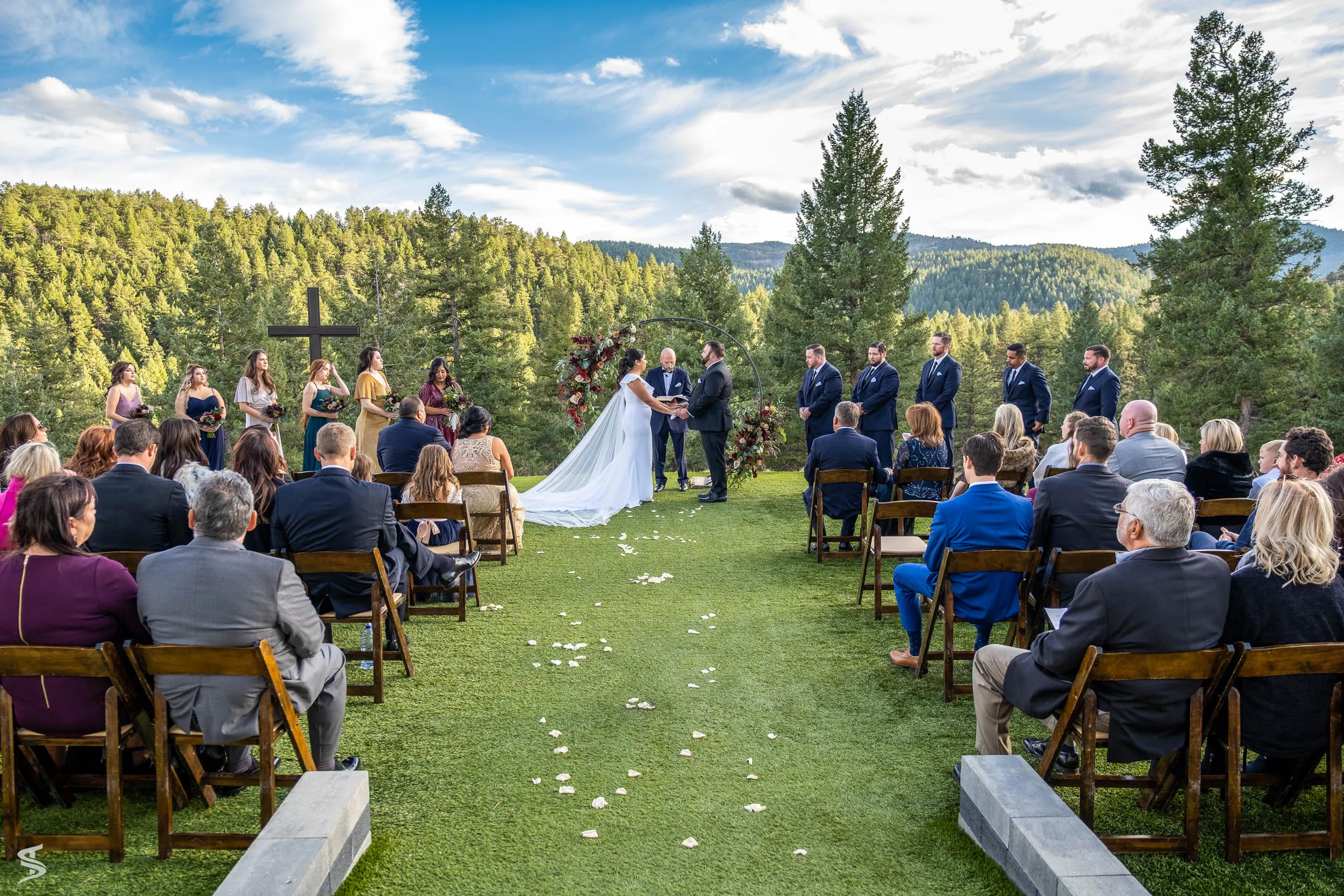 Guests watching an outdoor destination wedding ceremony on a lawn overlooking pine-covered hills.
