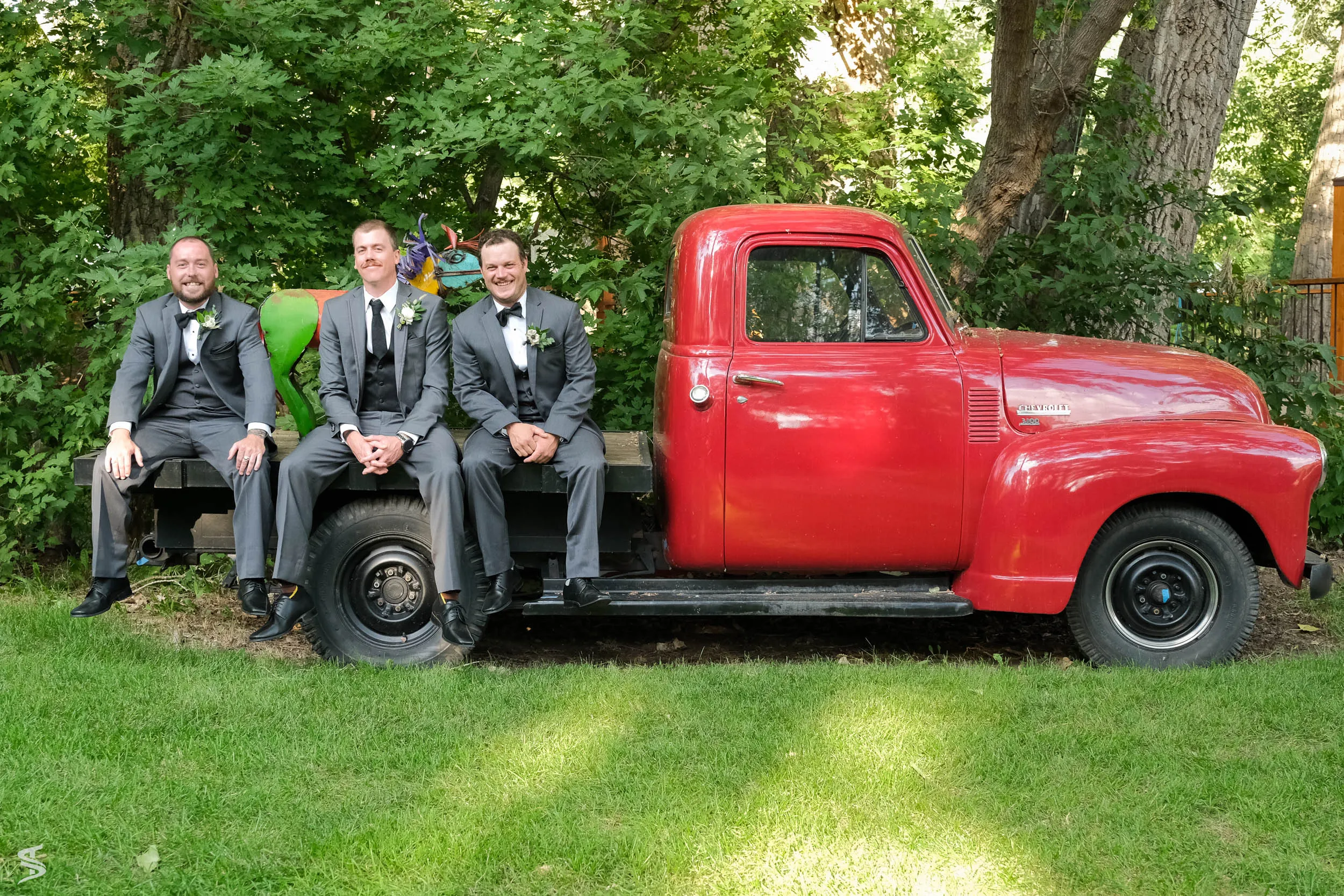 Three men in wedding suits sitting on the back of a vintage red pickup truck beneath leafy trees.