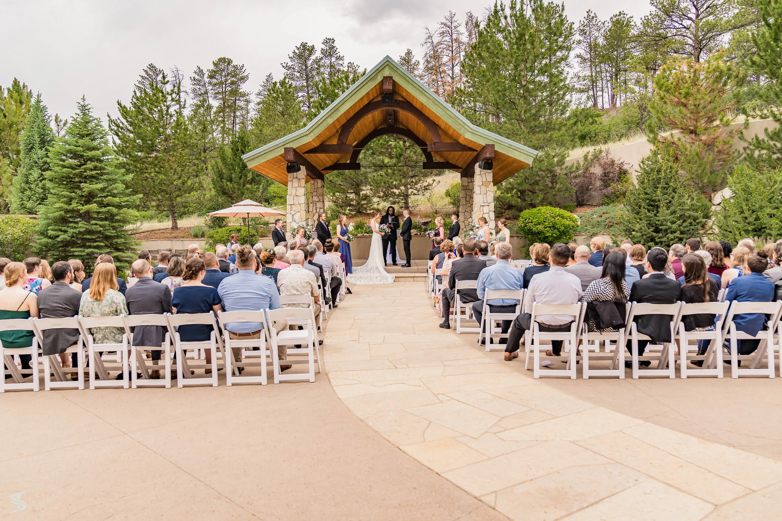 Guests seated along a stone aisle during an outdoor destination wedding ceremony beneath a timber pavilion.