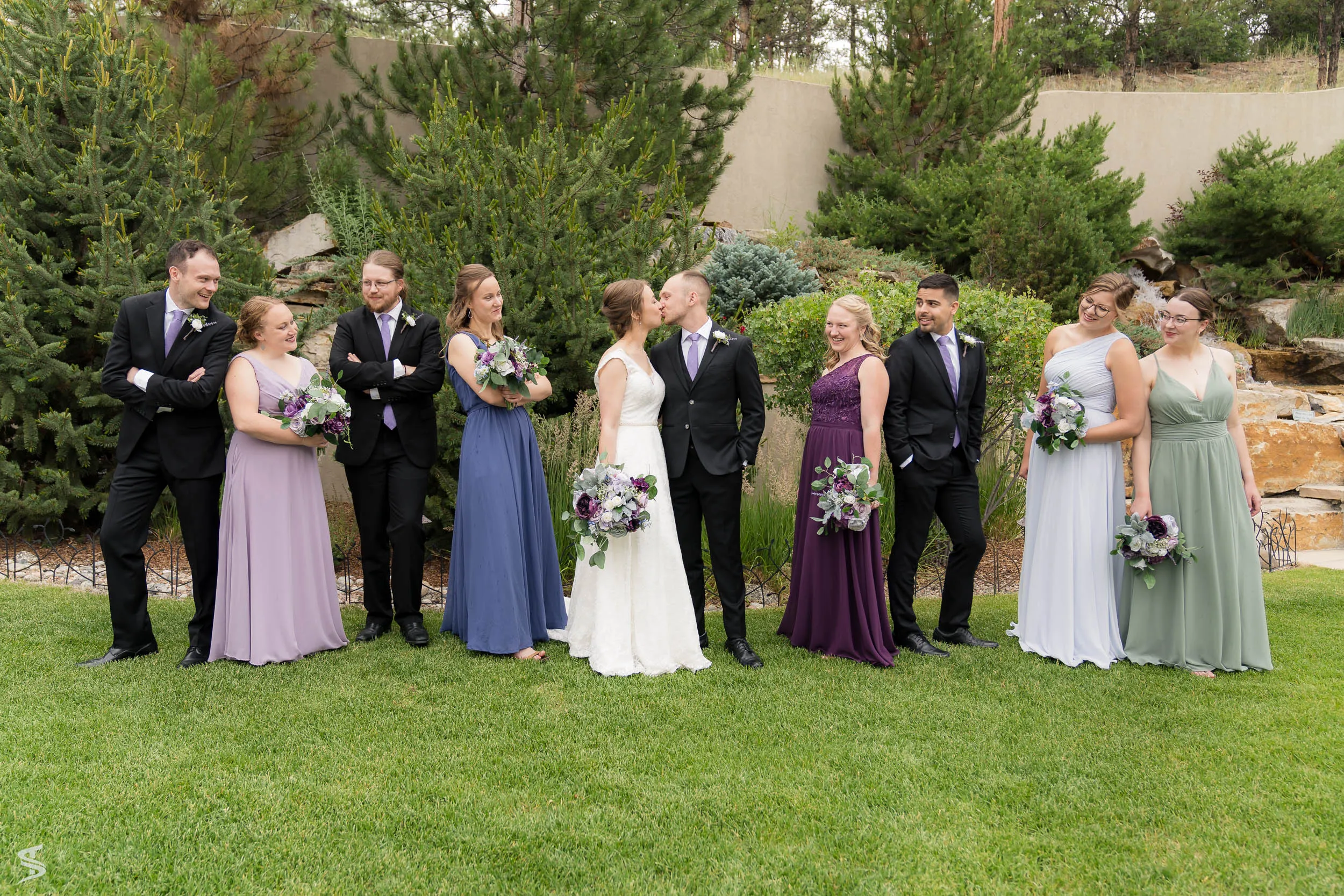 Bride, groom, and wedding party sharing a relaxed moment on a garden lawn beside a rock waterfall.