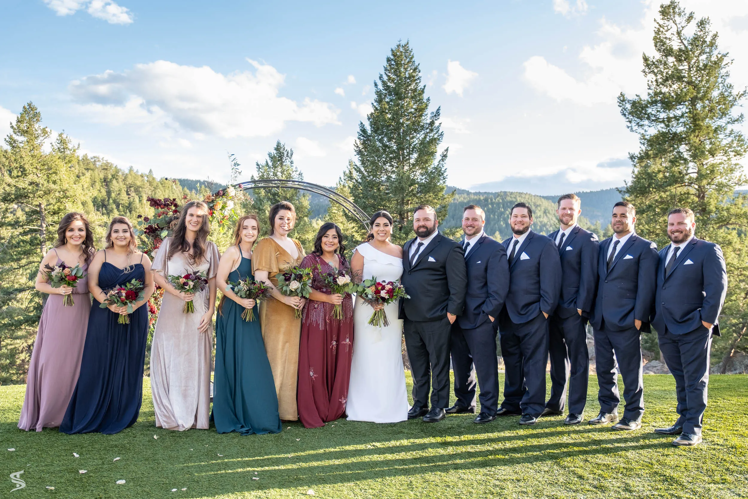 Bride, groom, and wedding party posing on a sunlit lawn with pine trees and mountains behind them.