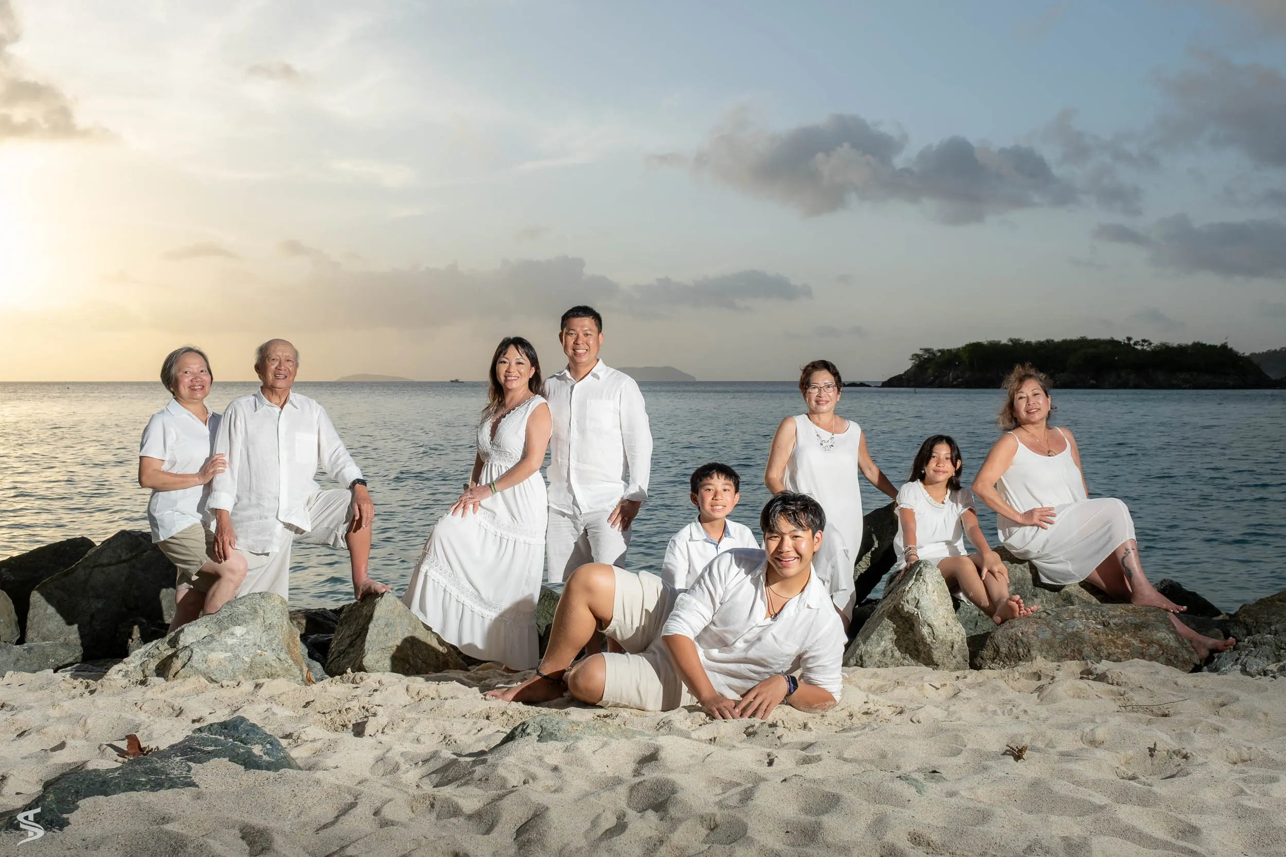 Family group in white outfits posing among shoreline rocks, with the sea and an island behind them.