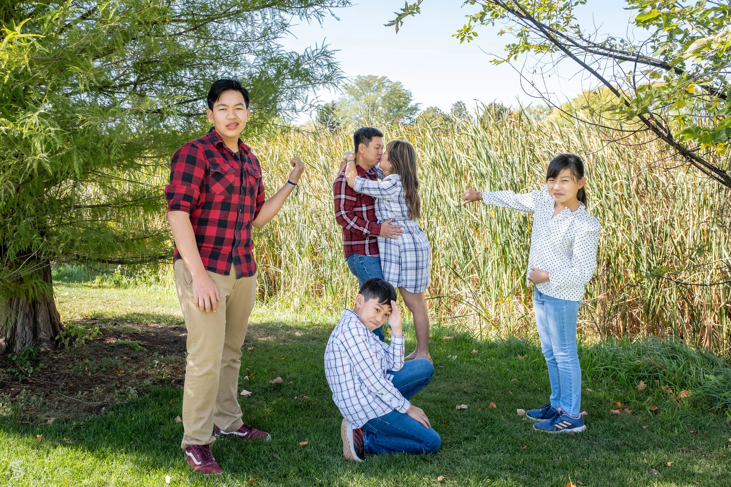Family of five striking playful poses beneath trees beside tall reeds.