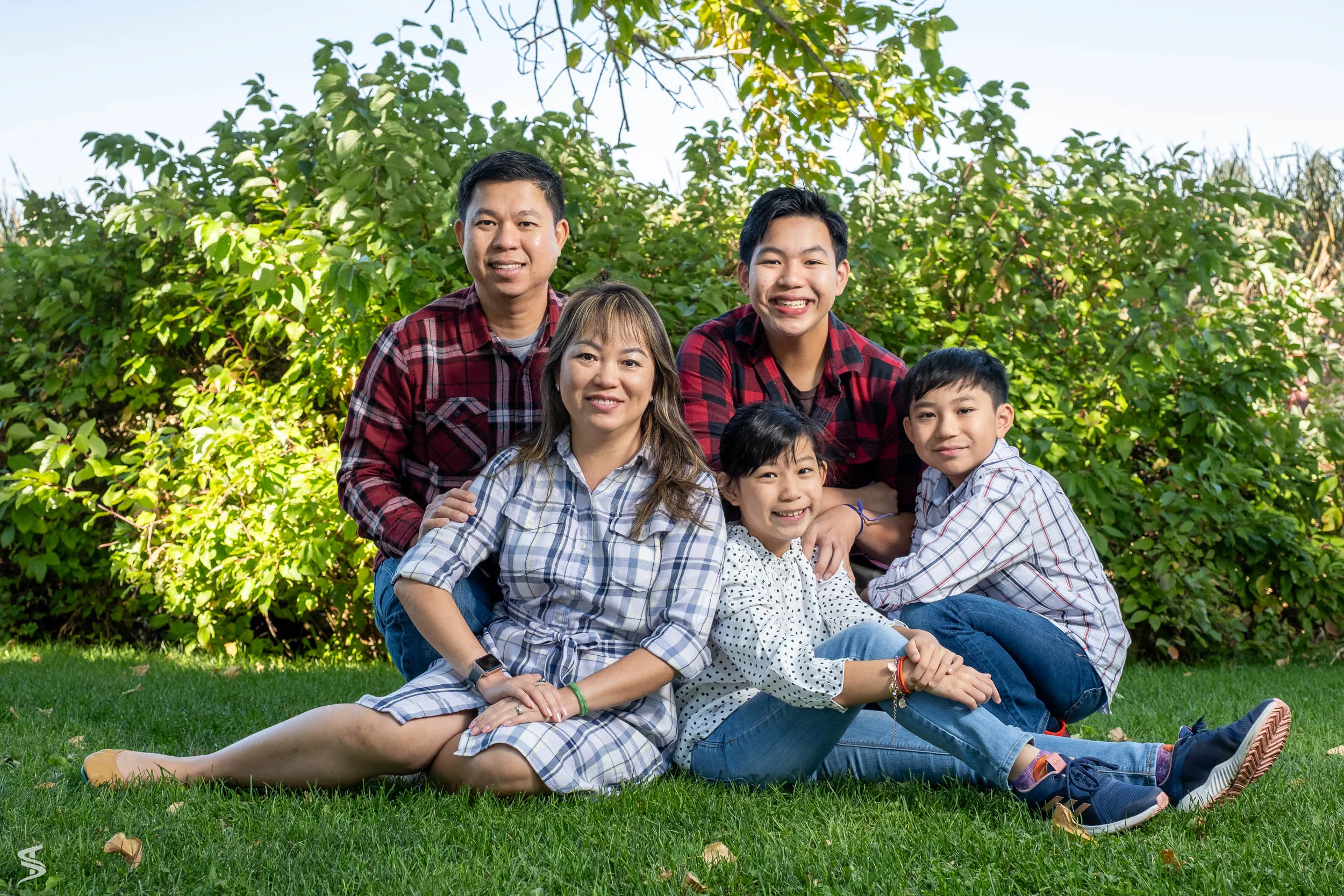 Family of five smiling together while seated on the grass in front of leafy shrubs.