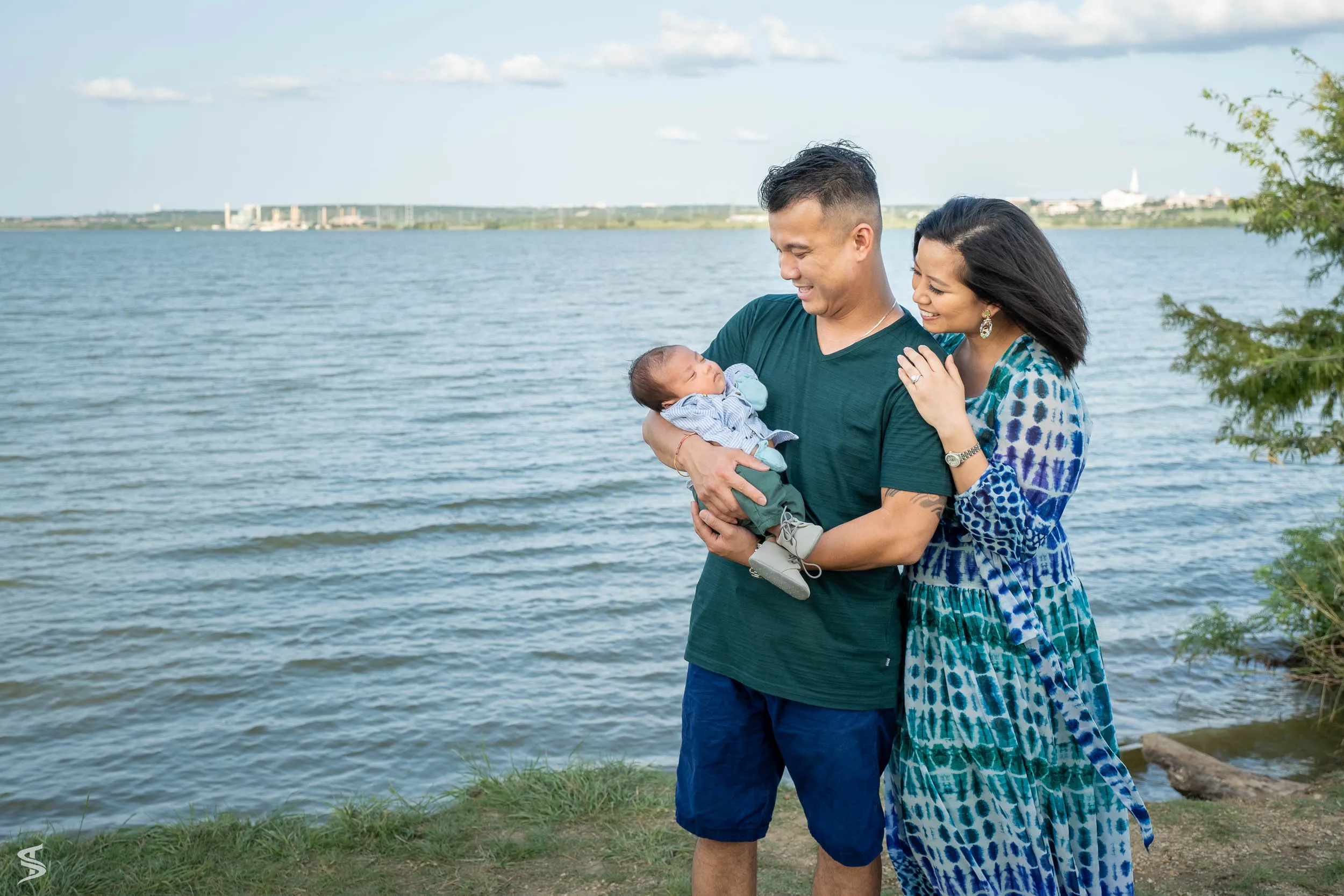 Parents looking down at their newborn baby while standing together beside a lake.