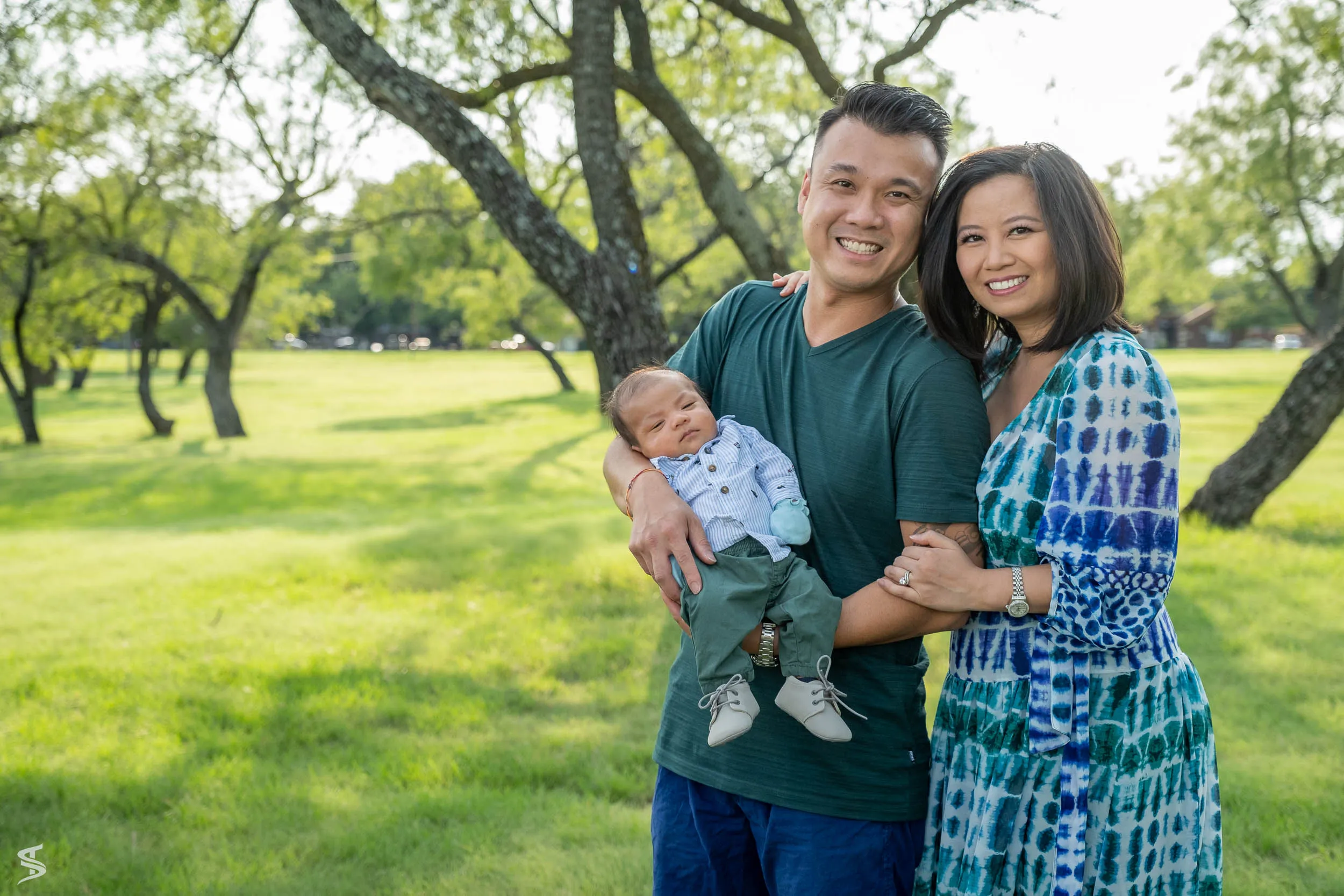 Smiling parents holding their newborn baby beneath leafy trees in a green park.