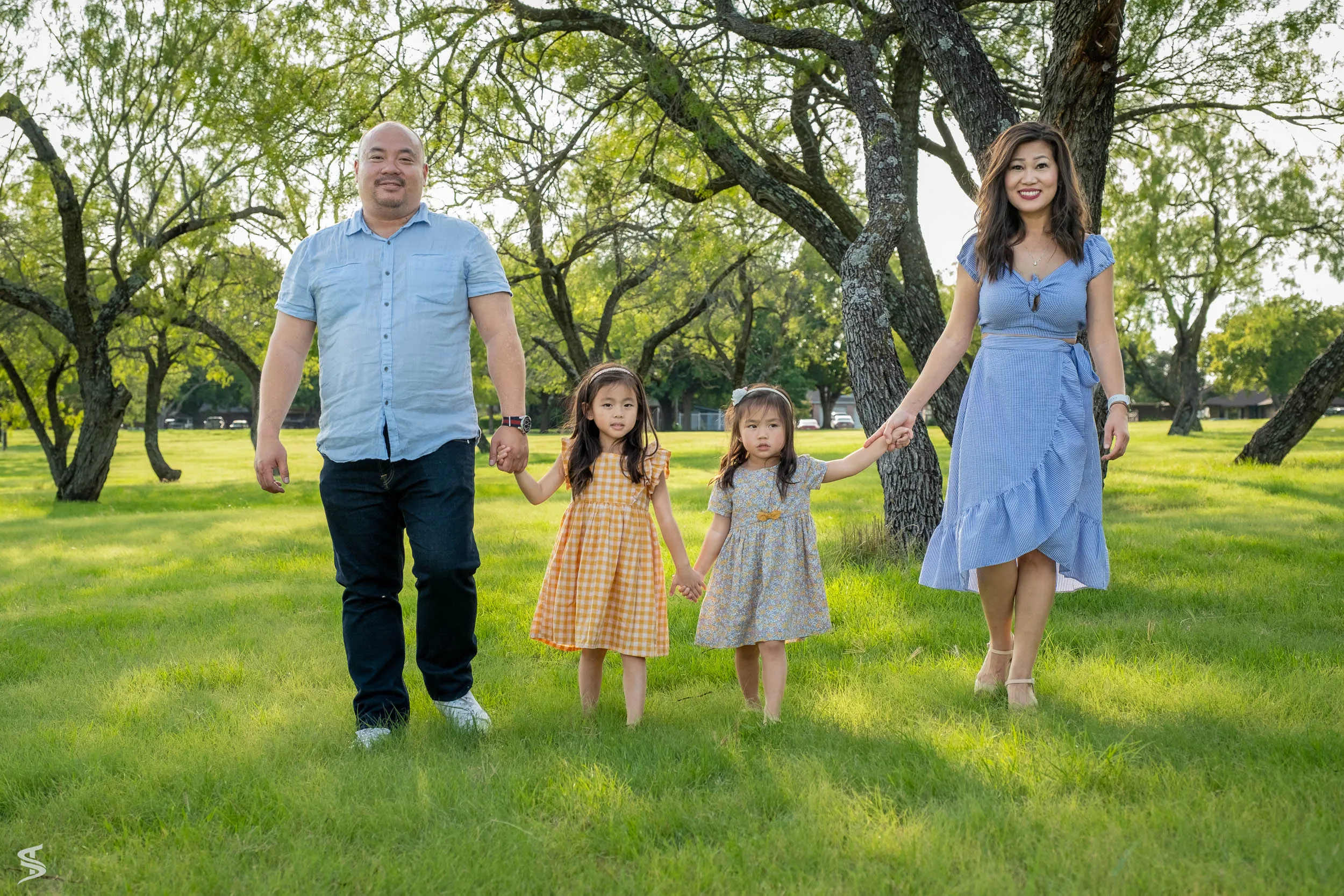 Parents walking hand in hand with their two young daughters through a sunlit park.