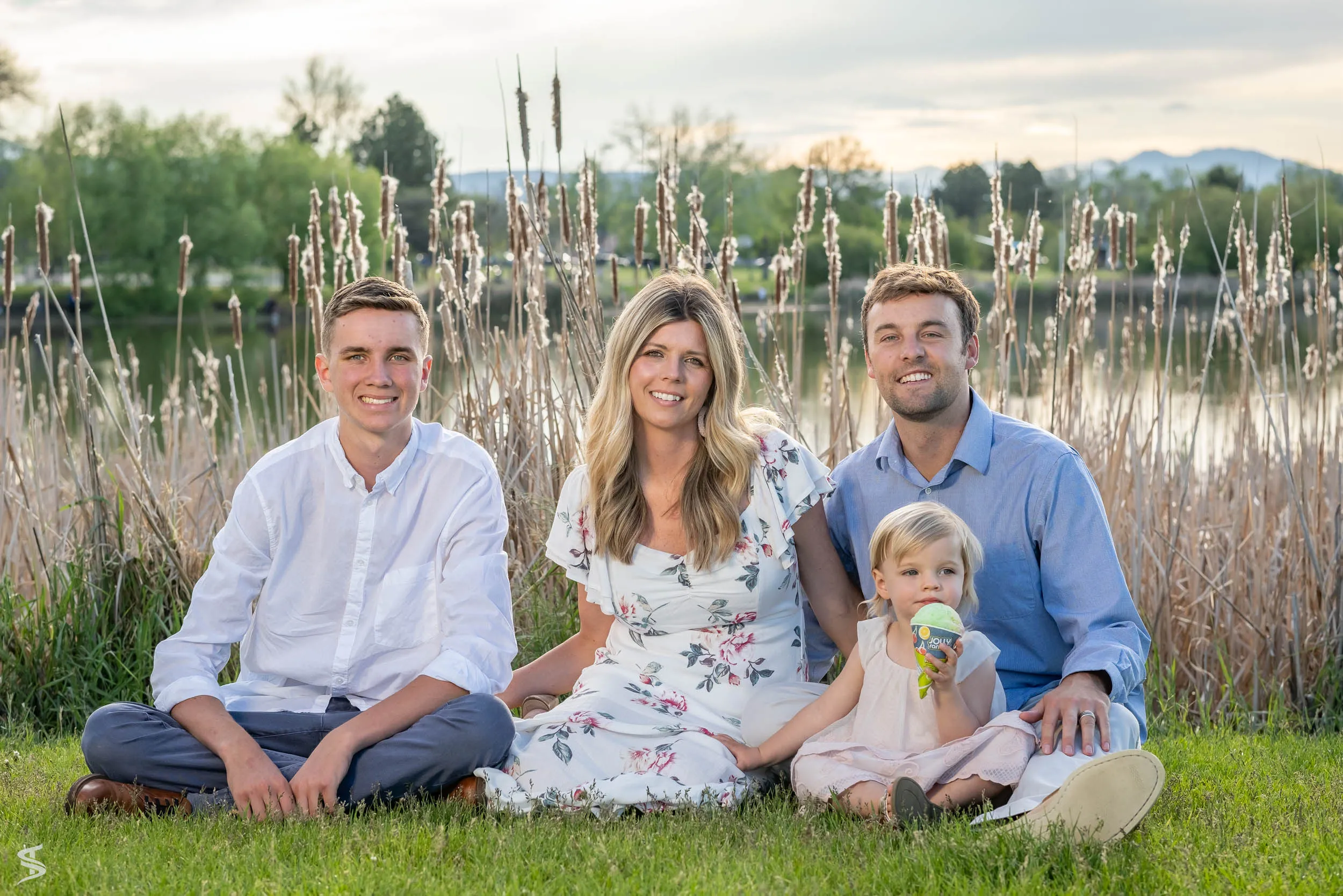 Three adults and a young child sitting together on the grass beside a lake at sunset.