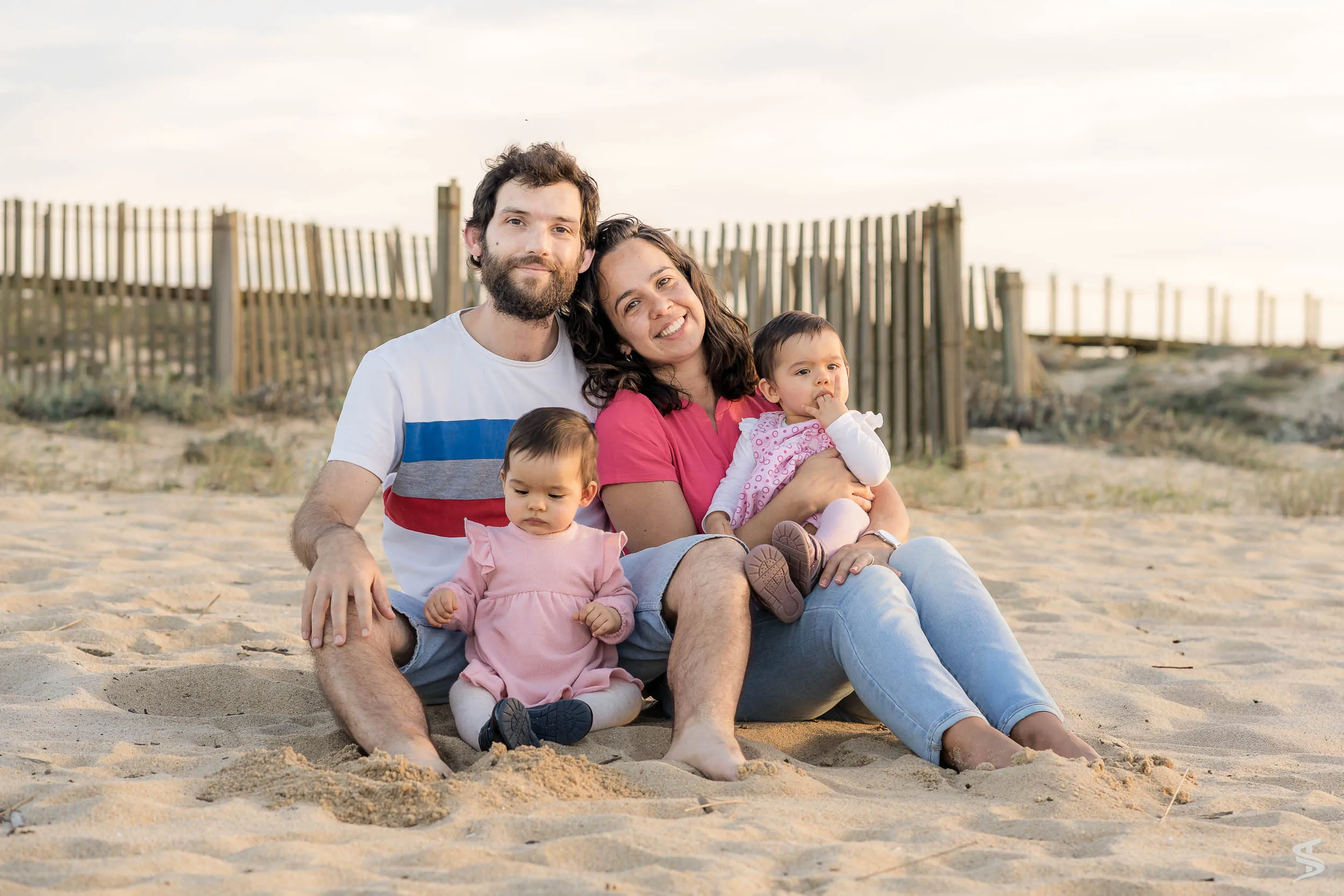 Parents and two young children sitting together on the sand beside a wooden beach fence.