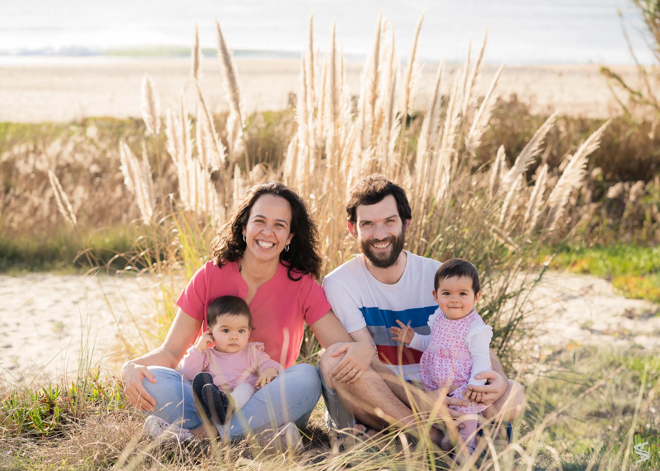 Parents sitting with their two young children among coastal grasses beside a sandy beach.