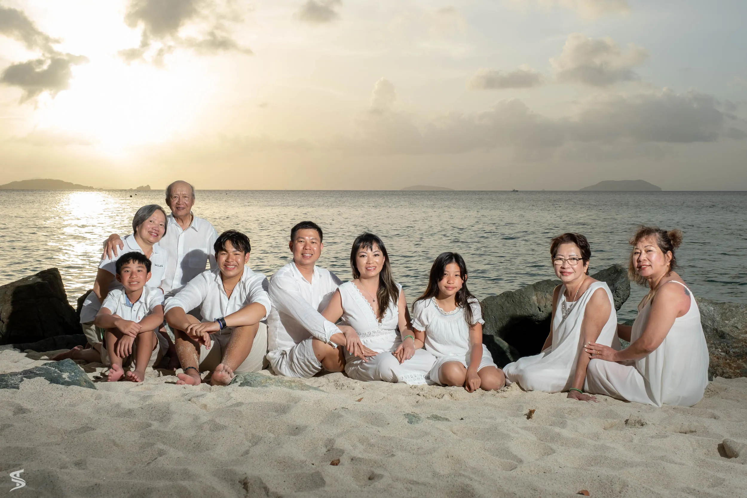 Family group in coordinated white outfits sitting together on a sandy beach at sunset.
