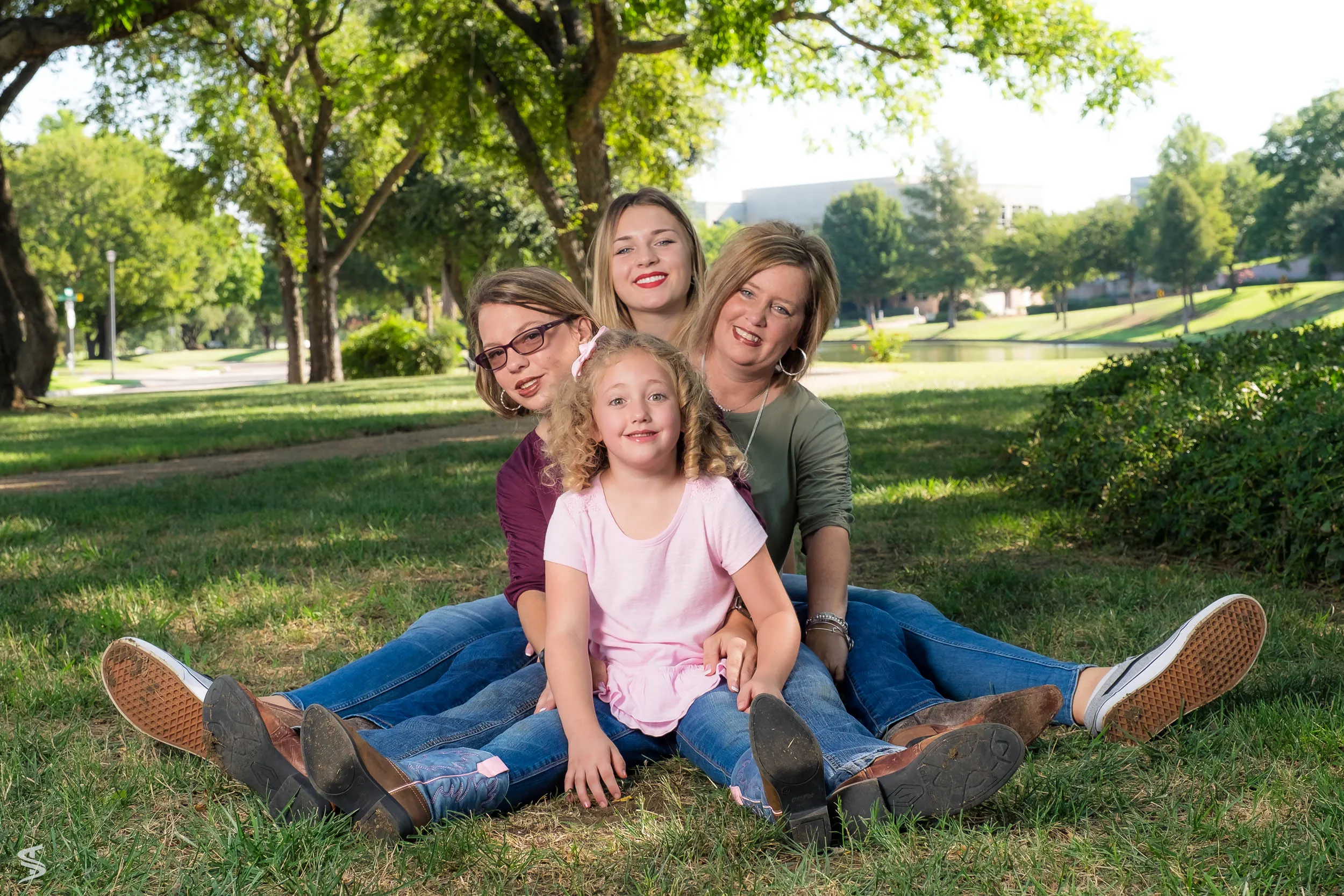 Woman and three children smiling together while seated on the grass in a sunlit park.